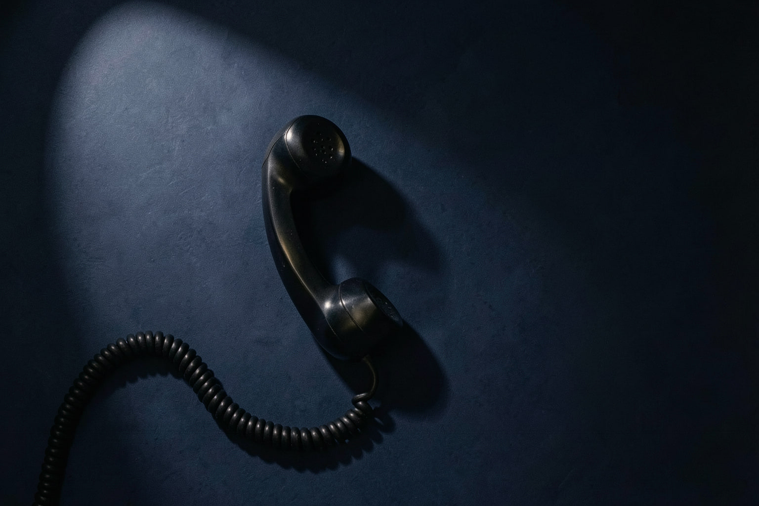 A black telephone receiver on its own, its coiled cord curling away under a single hard light.