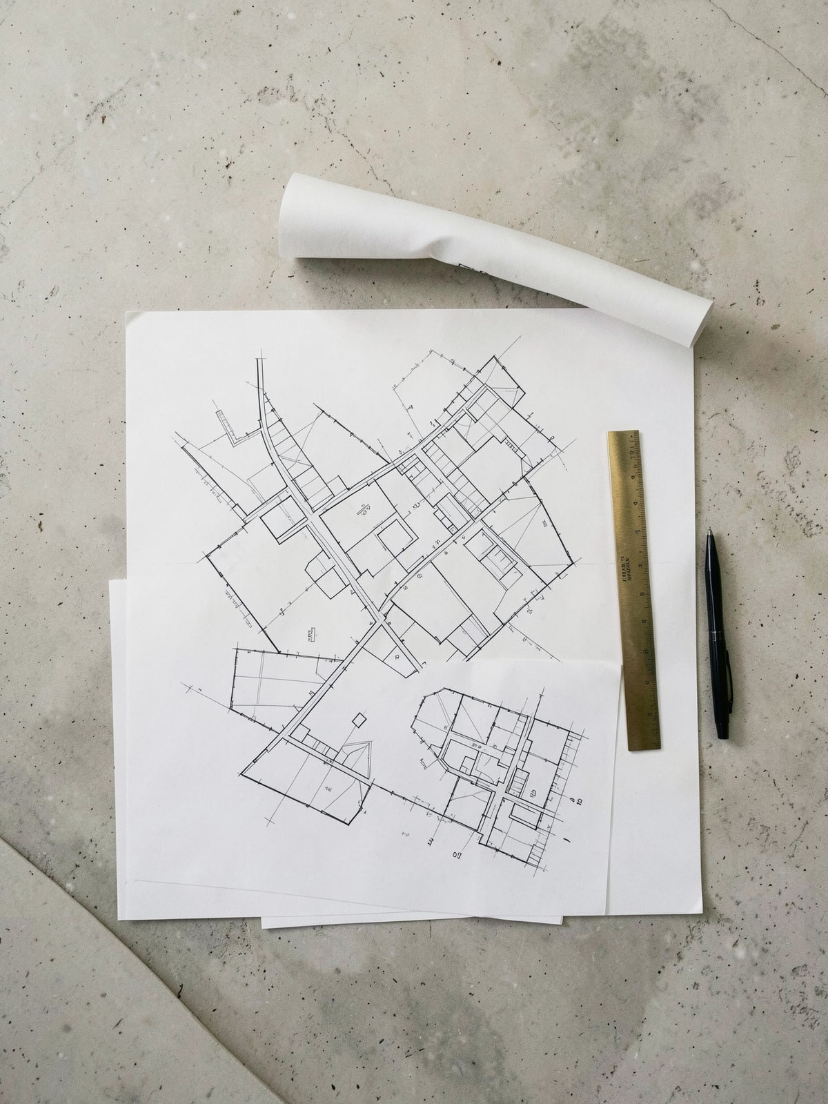Architectural site and floor plans laid out on a limestone table, a rolled drawing above them and a brass scale and pen alongside.